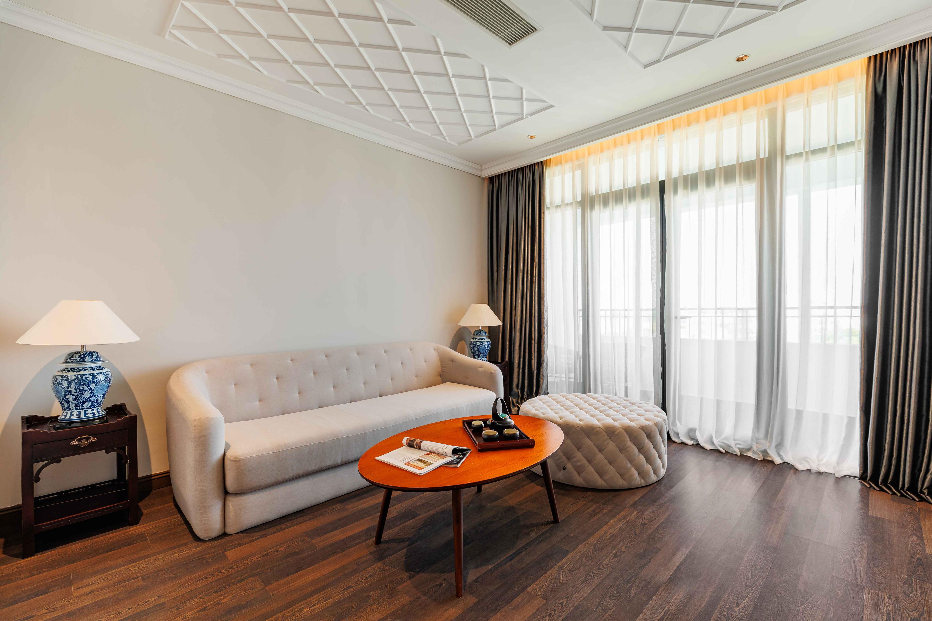 Spacious living area with a light-colored sofa and a round wooden coffee table. Two lamps provide soft lighting, and large windows with sheer curtains allow natural light to brighten the room. A cozy atmosphere is enhanced by a small decorative ottoman nearby