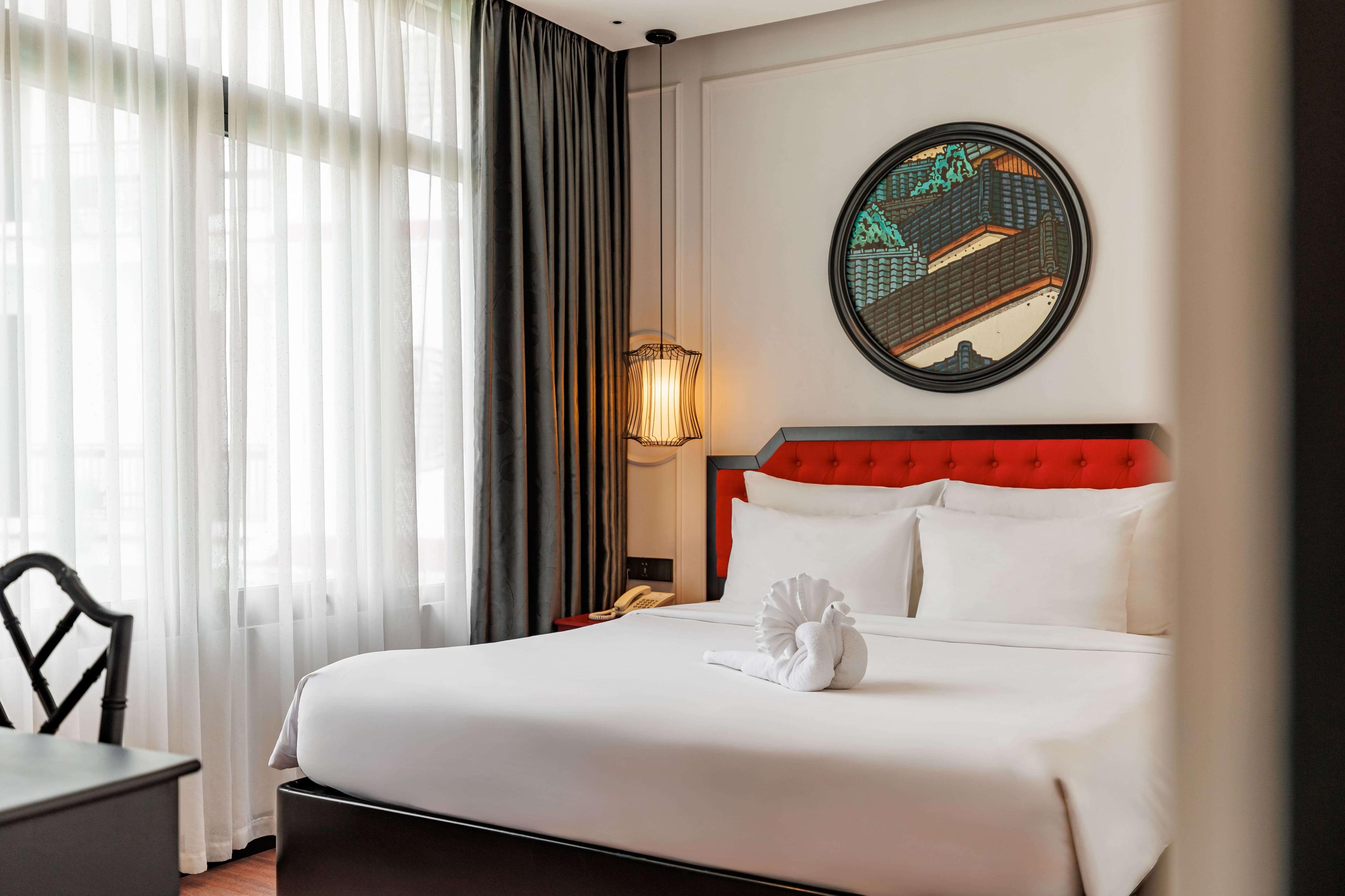 Inviting hotel room with a queen-sized bed dressed in white linens and a striking red headboard. Large windows draped with sheer curtains allow natural light, and a round mirror adds a decorative touch to the elegant decor